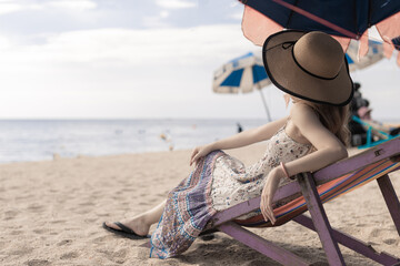 woman wearing wide brimmed hat and long dress relaxing on the beach chair