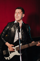 Fototapeta na wymiar Talented handsome young guitarist man singing a song in studio recording on red background surrounded by instruments. Passion, hobby, singer, electric guitar