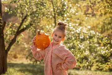 Happy smilling child girl in a knitted sweater is holding in hands ugly orange pumpkin. Deformed orange pumpkin with a damaged, ugly skin. Thanksgiving, harvest, halloween concept.