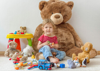 Sweet happy child boy having fun playing with his giant teddy bear and many colorful toys, indoor...