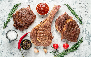 Variety of grilled beef steaks. Grilled tomahawk steak and ribeye steaks on a light stone background