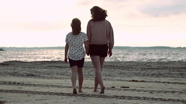 4K video of happy young mother walking holding hand with teen daughter on beach during sunset