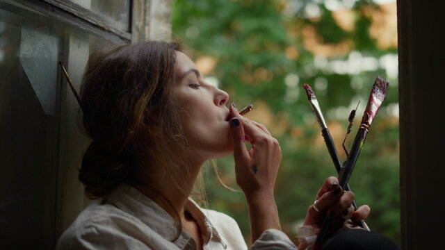 Creative woman holding cigarette indoors. Female painter smoking on windowsill.