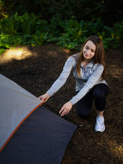 Young woman in sportswear lays out the tent. Road, dense green forest in the background. Clear sunny day. There is an empty space for your insert.