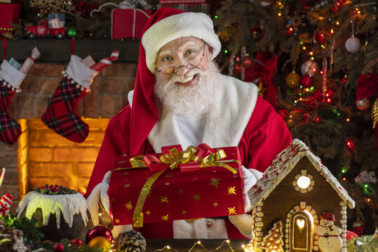 Santa Claus with gift box presents near fireplace and Christmas tree. Wooden house, New Year's cheerful mood Spirit of Christmas. Senior man with real white beard cosplay Father Christmas.