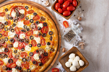 On a beautiful white napkin, we see pizza on a wooden plate. The pizza is beautifully decorated with olives, tomatoes, cheese. There are tomatoes and mushrooms in the bowls to the right of the pizza.
