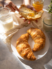In the photo we see croissants on a white earthenware plate. On the right in the photo there is milk in a glass glass. In the background is a croissant spread with jam. White silk tablecloth..