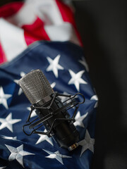 In the photo we see a microphone in the foreground, in the background is the American state flag. A symbol of freedom and patriotism. Dark gray background. Careful viewing. No people.