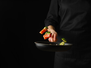 The cook is holding a frying pan with red fish, cut into pieces. Levitation. The cook in a black apron. The black background and the bright colors of the ingredients create contrast.