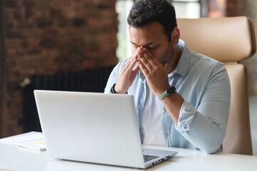 Fatigued indian man using laptop for working, a guy in casual wear is touching and massaging the...