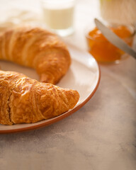 In the photo we see a morning breakfast - two croissants on a white earthenware plate, orange jam in a glass vase, a table knife. Light background. High angle view.