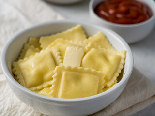 In the foreground, we see a white earthenware plate of ravioli. In the background is a deep bowl of sauce. White background. Careful viewing. High angle view. There are no people in the photo.