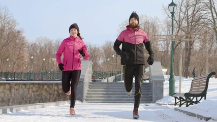 Slow motion cheerful couple wearing pink and black sportswear running in winter park. Tracking shot fit people enjoying outdoor activities together. Concept of sport