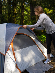 In the photo we see the process of setting up the tent. A woman hiker in the forest lays out a tent for rest. Sunny day. In the background we see a forest. The woman is wearing sportswear and shoes.