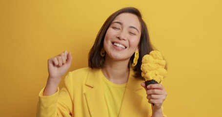 Positive young Asian woman with dark hair has fun dance to favorite music holds delicious ice cream expresses positive emotions feels upbeat dressed formally isolated over yellow background.