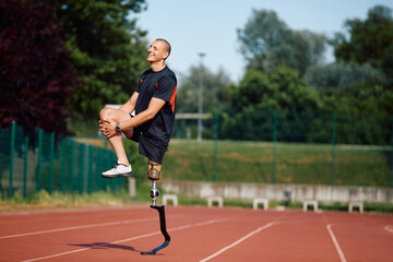 Happy disabled athlete stretching his leg while warming up for run on running track at the stadium.