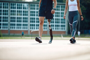 Unrecognizable athlete with leg prosthetic and his female friend walking at sports field while...