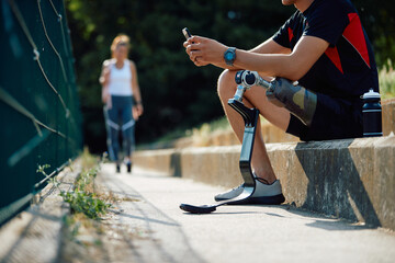 Unrecognizable sportsman with leg prosthetic uses smartphone while relaxing on staircase after...