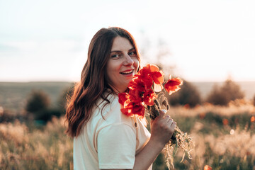 Caucasian Happy beautiful young girl with a poppy bouquet in her hands in a summer field at sunset. Tourism, traveling and healthy lifestyle concept.