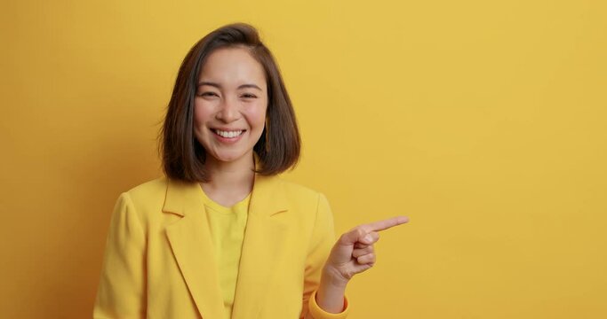 Smiling positive attractive young Asian lady in elegant clothes indicates away at blank space demonstrates product or company logo stands pleased against yellow background. Advertisement concept