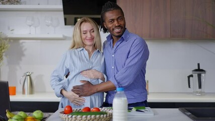 Happy interracial couple standing in kitchen caressing pregnant belly. Happy smiling African American man and Caucasian woman stroking tummy in slow motion - Powered by Adobe