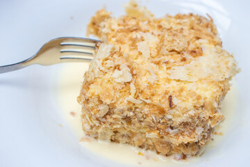 Piece of Napoleon tart on white plate with fork. Sweet dessert for breakfast. Vanilla creamy cake. French pie called Napoleon, close up. Homemade biscuit. Delicious dessert in summer cafe. 