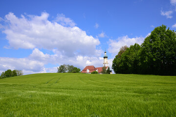 Landscape in Bavaria with rolling green hills against a blue sky with large clouds and a church on the hill
