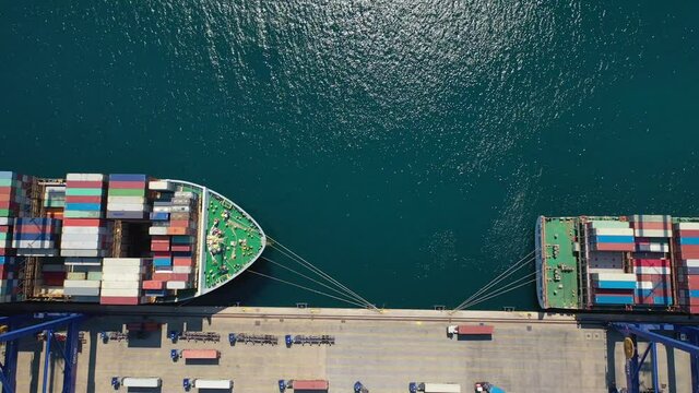 Aerial top down pan video of huge cranes load - unload colourful containers in anchored ship below in container terminal port