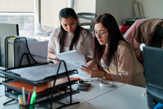 two young latina business women talking and working together in office, using documents and office supplies, teamwork concept