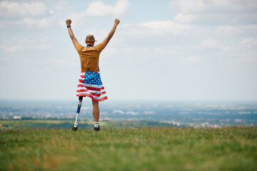 Rear view of disabled American man with leg prosthesis stands with raised arms on top of hill. Copy space.