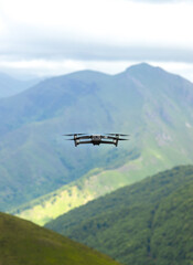 Fototapeta na wymiar Drone flies away over a mountainous landscape. The drone can be seen from behind flying away. In the background you can see a mountain and below it a forest and a green meadow.