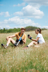 Happy man with leg disability and his female friend use smartphone and relax in grass while hiking in nature.