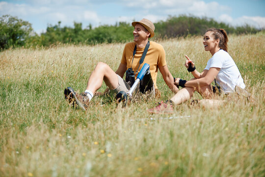 Happy hiker with leg disability has fun while relaxing in grass and talking with female friend.