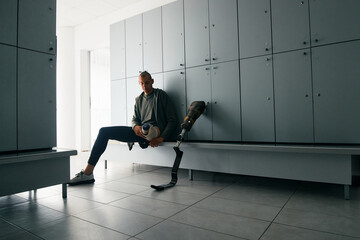 Athletic amputee putting on prosthetic leg in locker room and preparing for sports training at gym.