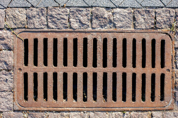 rusty manhole rectangular grating of the drainage system of the pedestrian sidewalk from rough cut stone tiles top view.