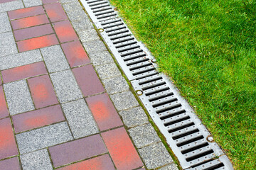 iron grating line of the drainage system of the pedestrian sidewalk made of stone tiles landscaping of the park with engineering systems on side of the path with the green lawn of the park, nobody.