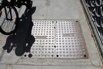 City telephone network hatch cover on the sidewalk. The shadow of a passing cyclist. A bright glare...