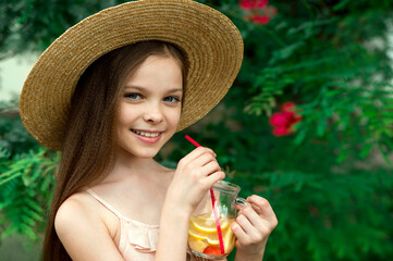 Cute little girl in summer hat drinks lemonade front of green leaves background. Kid holds glass...