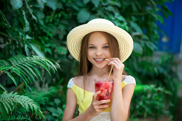 Cute little girl in summer hat drinks watermelon lemonade front of green leaves background. Kid holds glass with summer beverage cold tea.