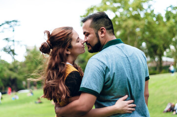 young hispanic latin couple of a white girl with red hair and a boy with a beard and short hair are hugging in a park.