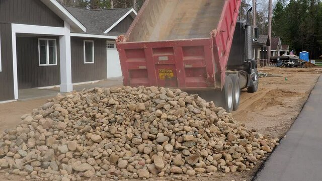 Dump truck drives forward and lowers its red, open box, after unloading a pile of landscape rocks onto the dirt. Dust rises and tailgate swings in this clip at new home construction site, with audio.