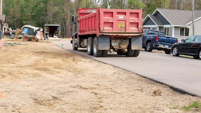 Red dump truck drives on asphalt pavement street at a new home construction site.