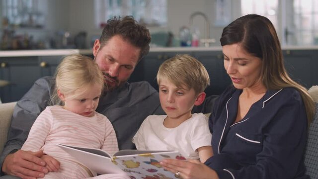 Family in pyjamas having fun sitting on sofa reading bedtime story from book together - shot in slow motion 