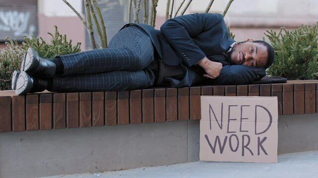 Lonely unhappy upset African American guy who lost job lying on park bench. Black millennial has forfeit income due coronavirus. Young businessman holding cardboard sign need job waiting for offers