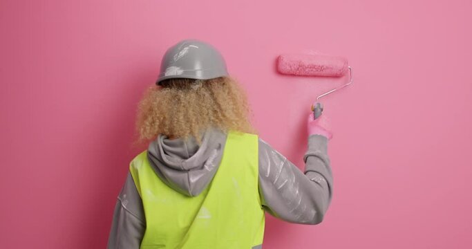 Back view of busy female painter does repair of apartment paints wall in pink uses painting roller wears hardhat and uniform stands faceless. Maintenance remodeling and refurbishment concept