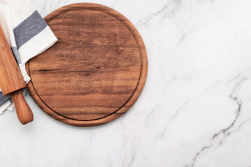 Empty wooden pizza platter with napkin and rolling pin set up on marble stone kitchen table. Pizza board and tablecloth on white marble background.