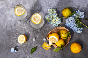 Water with lemon in glasses, fruits and flowers on a gray concrete background.