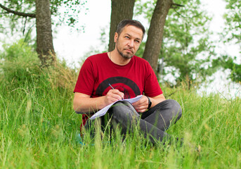 Naklejka na ściany i meble A man in the park, sitting in the grass and writing with a pen in an agenda.