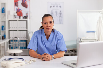 Portrait of pretty medical nurse smiling at camera in hospital office wearing blue uniform. Healthcare practitioner sitting at desk using computer in modern clinic looking at monitor, medicine.