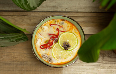 Top view of a bowl of tom yam soup surrounded by green plant leaves. Shrimps, mushrooms, chili slices, lime slices and baby octopus in a bowl on a wooden board.